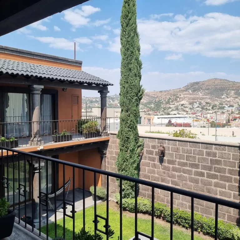 A balcony at Villa Serena overlooking a lush green courtyard, a tall cypress tree, and distant mountains. - Villa Serena