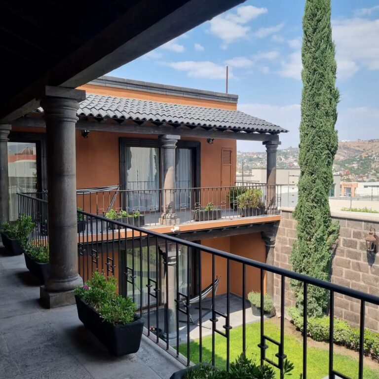 A balcony at Villa Serena overlooking a lush green courtyard, a tall cypress tree, and distant mountains. - Villa Serena