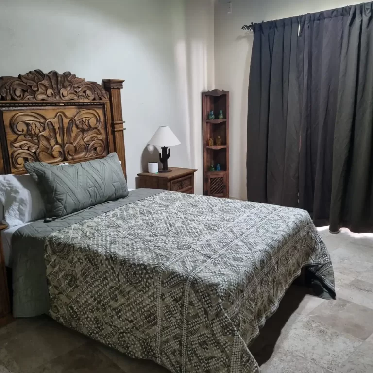 A bedroom at Villa Serena featuring a double bed with a patterned quilt, a wooden headboard, a nightstand with a lamp, and dark curtains. - Villa Serena