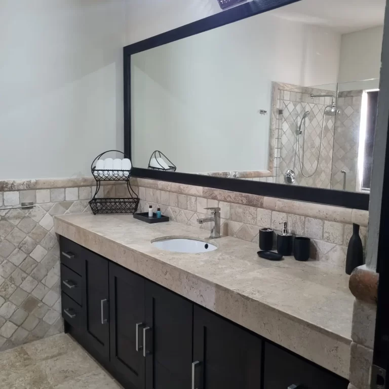 A modern bathroom at Villa Serena featuring a double sink with a marble countertop, dark cabinets, a large mirror, and a shower stall visible in the background. - Villa Serena