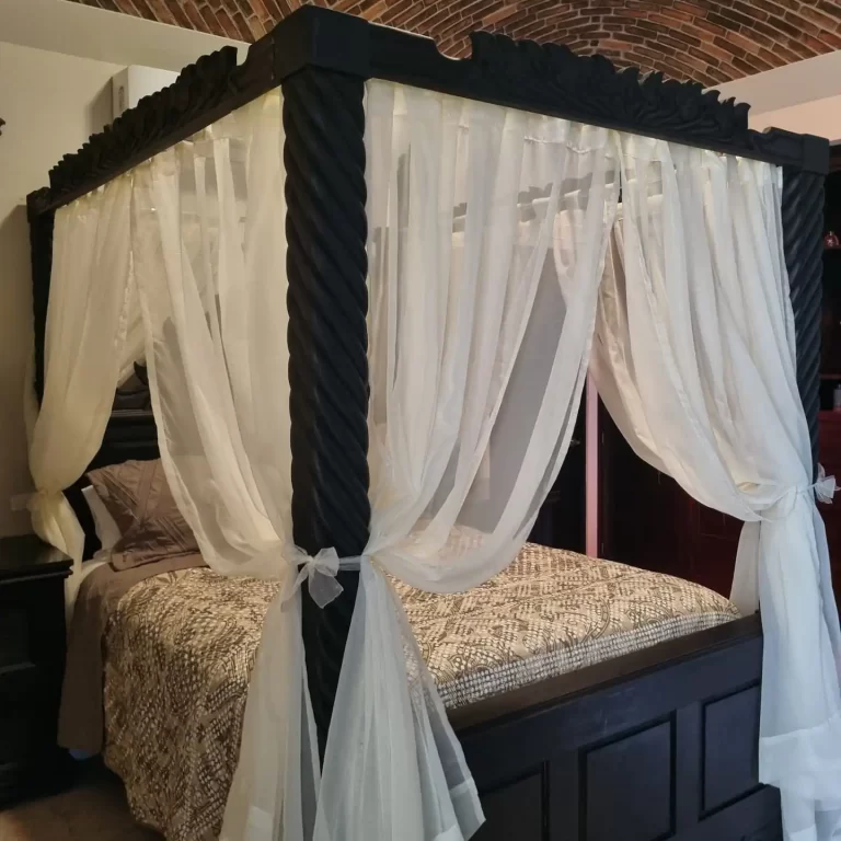 A four-poster bed with a carved wooden headboard and sheer white curtains. The bed is made with brown bedding.